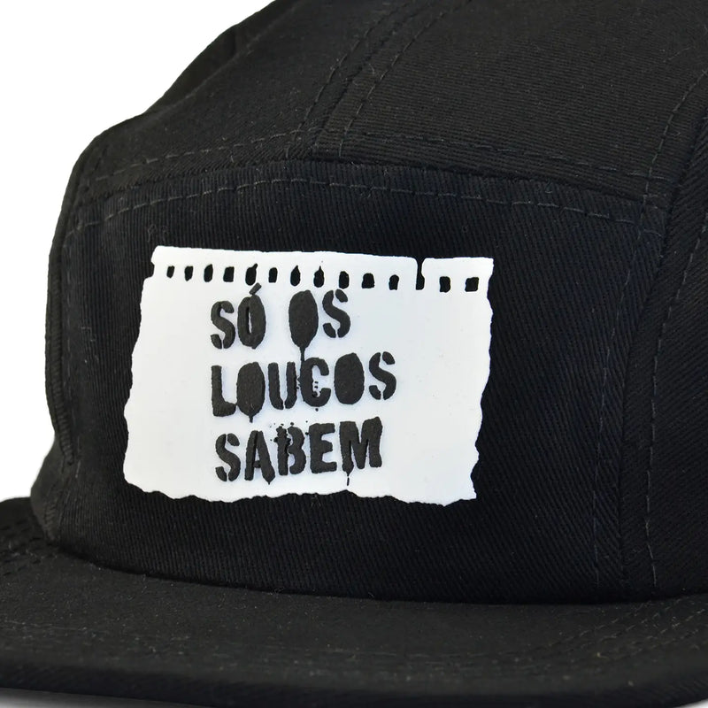 Boné five panel preto com patch branco em forma de papel rasgado e frase "Só os loucos sabem" em destaque na frente.