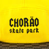 Boné five panel amarelo com aba xadrez marrom e bege, frase CHORÃO skate park centralizada em preto.