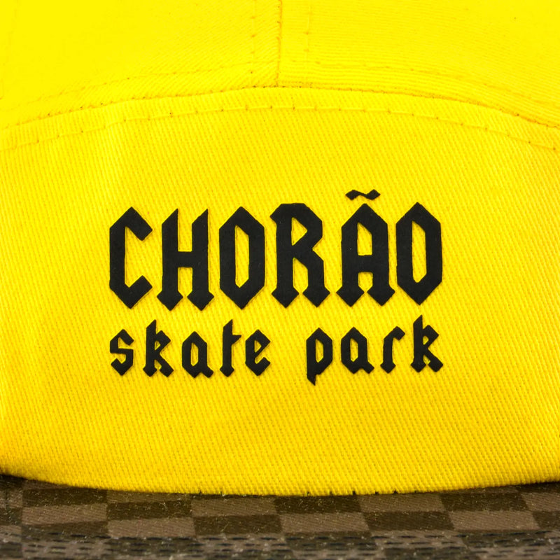 Boné five panel amarelo com aba xadrez marrom e bege, frase CHORÃO skate park centralizada em preto.